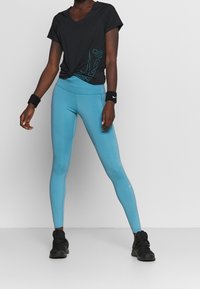Persona con una camiseta negra de mangas cortas, leggings azules de talle alto con perforaciones cerca de los tobillos, zapatillas negras y pulseras negras.