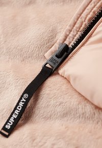 Giacca in finta pelliccia di un morbido color pesca, con una zip nera dotata di un tirante con il marchio "SUPERDRY", in contrasto con il tessuto testurizzato.