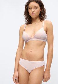 Νεαρή γυναίκα με κατσαρά μαλλιά, φορώντας ροζ ανοιχτό σουτιέν και ταιριαστό εσώρουχο μπροστά σε απλό ανοιχτό φωτεινό φόντο.