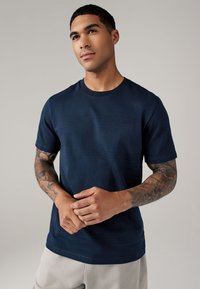 Navyblauwe t-shirt met korte mouwen, horizontale ribbelstructuur, ronde hals en een relaxte pasvorm, gedragen door een persoon met tatoeages op hun armen.