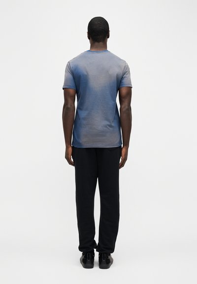 Άνδρας που στέκεται γυρισμένος την πλάτη, φοράει ένα ξεθωριασμένο μπλε πουκάμισο με κοντά μανίκια, μαύρο παντελόνι και μαύρα αθλητικά παπούτσια, μπροστά σε απλό λευκό φόντο.