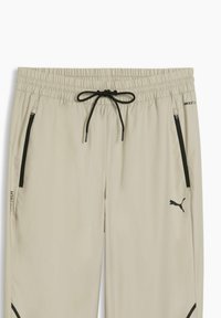 Pantalon de sport beige clair en matière lisse et légère. Comprend une taille élastique avec cordon noir, des poches zippées et des accents noirs.