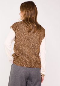 Maglione senza maniche tricot marrone con un design testurizzato e orlo a costine, abbinato a una camicia a maniche lunghe color crema e pantaloni grigi.