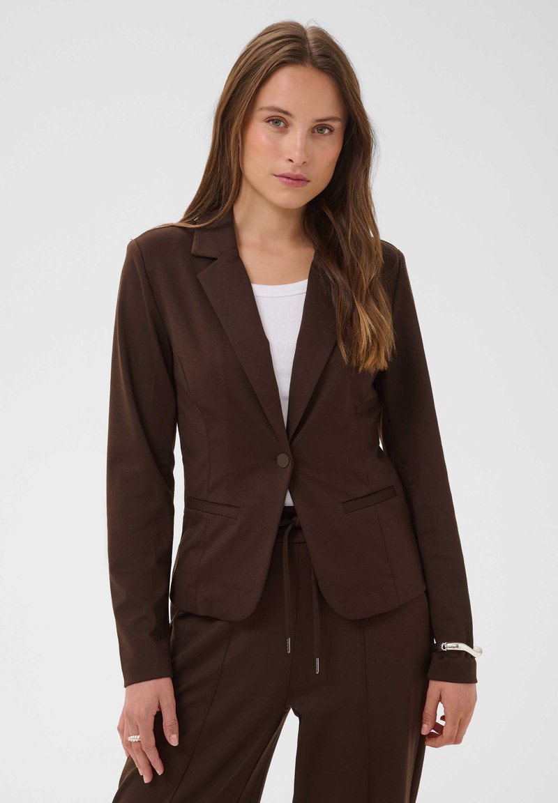 Femme portant un blazer marron foncé ajusté et un pantalon assorti sur un haut blanc, debout devant un fond clair uni.