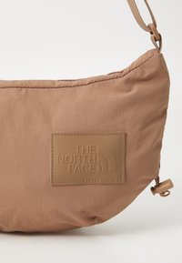 Bolso de hombro de nylon marrón con un parche de logo texturizado y rubberizado. Forma suave y curvada con una correa ajustable y un cierre deslizante.