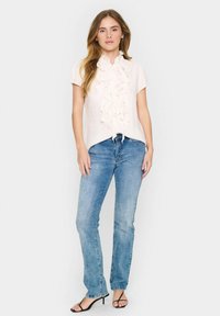 Lichtroze blouse met ruffle-voorkant en korte mouwen, met een strikdetail aan de voorkant, gedragen met blauwe spijkerbroek en zwarte hak-sandalen.