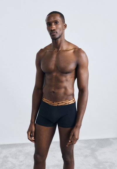 Schwarze Boxershorts mit einem orangen Bund, der handschriftlichen Text zeigt. Modell mit durchtrainierter Figur steht vor einem hellen Hintergrund.