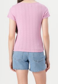 Femme portant un haut texturé violet clair à manches courtes et un short en denim bleu clair, vue de dos sur fond blanc.