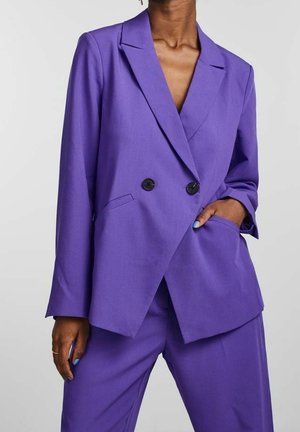 Person iført en lilla dobbeltradet blazer med sorte knapper og matchende bukser, den ene hånd i lommen og den anden ved siden.