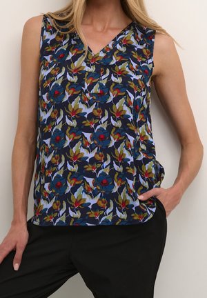 Haut sans manches bleu marine avec un motif floral multicolore porté par une personne aux cheveux blonds, assorti à un pantalon noir et main dans la poche.