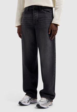 Persoon die losse zwarte spijkerbroek, wit shirt met lange mouwen en zilveren sportschoenen draagt, staand op een lichte achtergrond.