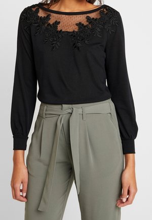 Haut noir à manches longues avec encolure transparente et détails en dentelle florale, associé à un pantalon taille haute vert olive avec ceinture en tissu nouée.