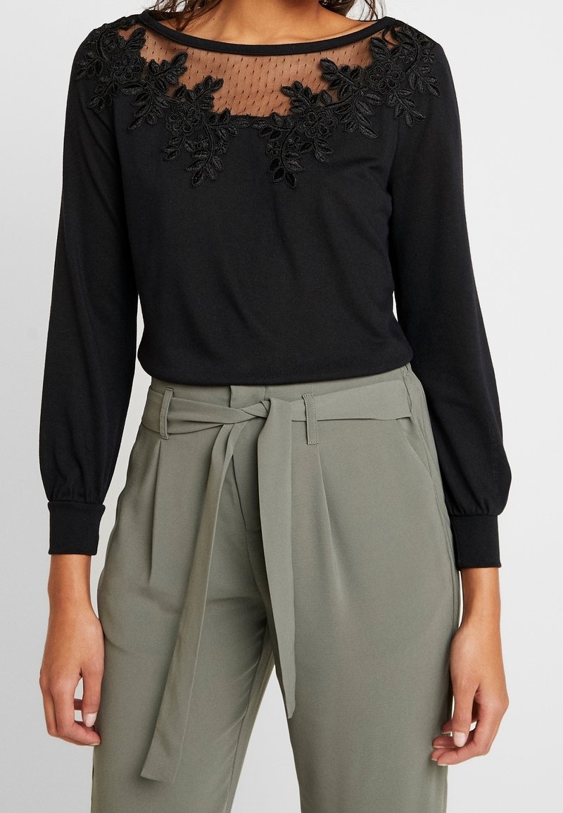 Haut noir à manches longues avec encolure transparente et détails en dentelle florale, associé à un pantalon taille haute vert olive avec ceinture en tissu nouée.