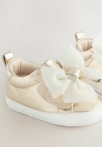 Goldene metallische Schuhe für Kleinkinder mit einer großen weißen Schleife, strukturiertem Material, glatter Oberfläche und weißer Gummisohle. Ergänzt durch kleine Fersenschlaufen.