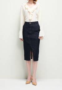 Frau mit einer cremefarbenen Rüschbluse, einem dunkelblauen Midirock mit Schlitz vorne und beigen High Heels, die vor einem schlichten Hintergrund steht.