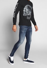 Camisa negra de manga larga con un diseño gráfico de calavera. Lleva puesto unos jeans de mezclilla azul ajustados y zapatillas azul marino con acentos blancos.