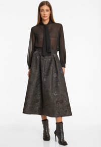 Vrouw gekleed in een doorschijnende zwarte blouse met strik, een metallic patroon midi rok en zwarte hoge enkelboots, staand tegen een witte achtergrond.