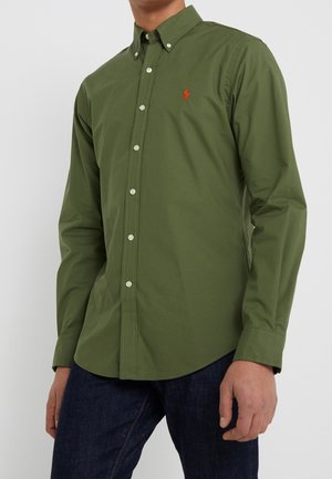 Uomo che indossa una camicia verde oliva a maniche lunghe abbottonata con colletto e jeans blu scuro su uno sfondo chiaro uniforme.