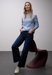 Femme en pull bleu clair et jean foncé appuyée sur une chaise moderne bordeaux sur un fond gris uni.