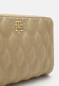 Beige bőr kluccs, steppelt mintával, arany színű logóval ellátott fém részekkel. Cipzáras záródás a tetején és texturált felület.