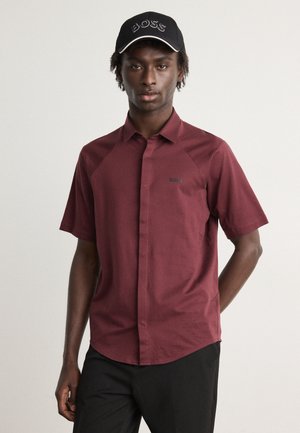 Camicia a maniche corte color bordeaux con colletto button-down, caratterizzata da un logo discreto sul lato sinistro. Indossata con un cappellino nero, logo ricamato e dettagli bianchi.