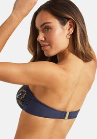 Navy bandeau top met geborduurde turquoise accenten, gouden hardware en zachte stof. Het ontwerp bevat een achterklep voor een veilige pasvorm.