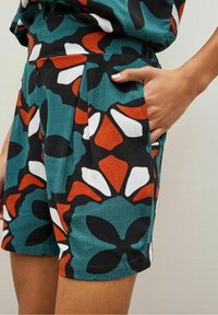 Personne portant un short à motifs floraux teal, noir, blanc et orange, avec une main partiellement dans la poche, sur un fond uni.
