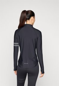Black long-sleeve athletic shirt with a high collar, textured grey stripes on the left arm, and a curved hem with decorative dots at the back.