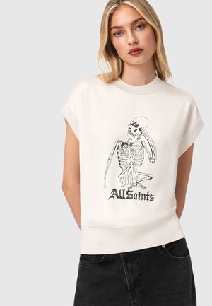 Blonde vrouw die een wit mouwloos top met een zwarte skelet dat een persoon vasthoudt en de tekst "All Saints" draagt, gecombineerd met een zwarte spijkerbroek.