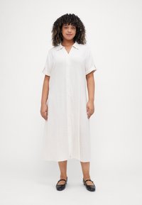 Short-sleeve button-up dress in light cream fabric with a relaxed fit, reaching mid-calf, paired with black flat shoes.