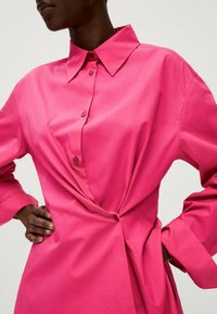 Nahaufnahme einer Person, die ein leuchtend pinkes Langarmhemdkleid mit einem Knoten-Detail an der Taille und einem großen spitzen Kragen trägt.