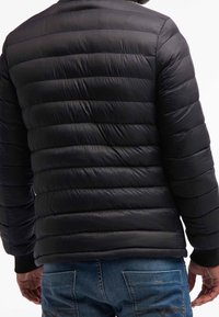 Giacca a vento nera con design trapuntato, caratterizzata da cuciture orizzontali e polsini a coste, indossata sopra jeans in denim blu.