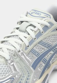 Gros plan d'une basket Asics blanche et bleu clair avec tissu en mesh, surpiqûres à motifs et lacets blancs noués en nœud.