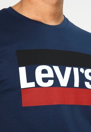 Man draagt een blauw T-shirt met het Levi’s-logo in grote witte letters, omlijst door zwarte en rode rechthoekige blokken.