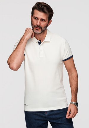 Uomo con barba che indossa una polo bianca con bordatura blu navy e jeans scuri, toccandosi il collo con la mano sinistra, in piedi davanti a uno sfondo uniforme.