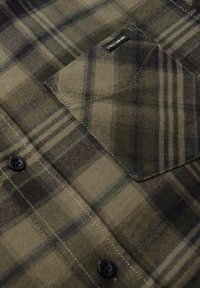 Camisa de franela verde olivo con un patrón de cuadros, botones negros y un bolsillo con parche que lleva una etiqueta. Textura suave y detalles de costura notables.