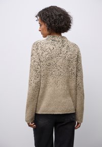 Beżowy sweter o teksturowanej powierzchni z puszystym wykończeniem, z wzorem w kropki, długimi rękawami i wysokim kołnierzem. Noszony z ciemnoniebieskimi dżinsami.