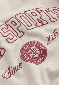 Gråt stof med fed bordeaux broderi med ordet "SPORTS" og en cirkulær logo. Detaljerne inkluderer "2023" og "Since" i kursiv skrift.
