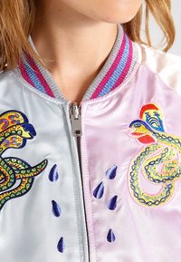 Kobieta nosząca pastelowy różowy i jasnoniebieski satynowy kurtkę z kolorowymi haftowanymi wzorami węży i niebieskimi kroplami wody.