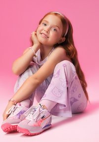 Puma sportbačiai pastelinėmis rožinėmis ir baltomis spalvomis su gradientiniu raštu. Mergaitė vilki šviesiai purpurines kelnes su Hello Kitty raštu ir aksesuarais. Rožinis fono spalva.