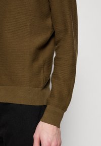 Homme portant un pull en maille marron texturé avec poignets et ourlet côtelés, associé à un pantalon noir, visible du milieu du torse à la main.