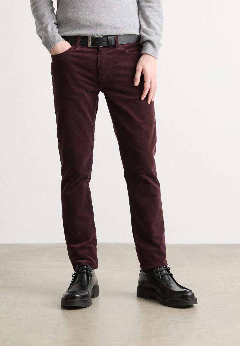 Pantalon en velours côtelé bordeaux à coupe slim, associé à des chaussures noires. Un pull gris complète le look, mettant en valeur le contraste des couleurs de la tenue.