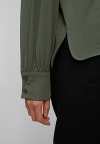 Blusa verde oliva con maniche lunghe arricciate e polsini con bottoni, abbinata a pantaloni neri. Tessuto liscio con una morbida texture.