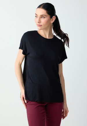 Femme avec une queue de cheval foncée, portant des boucles d'oreilles créoles dorées, un haut noir à manches courtes et un pantalon bordeaux, regardant vers sa gauche sur un fond blanc.