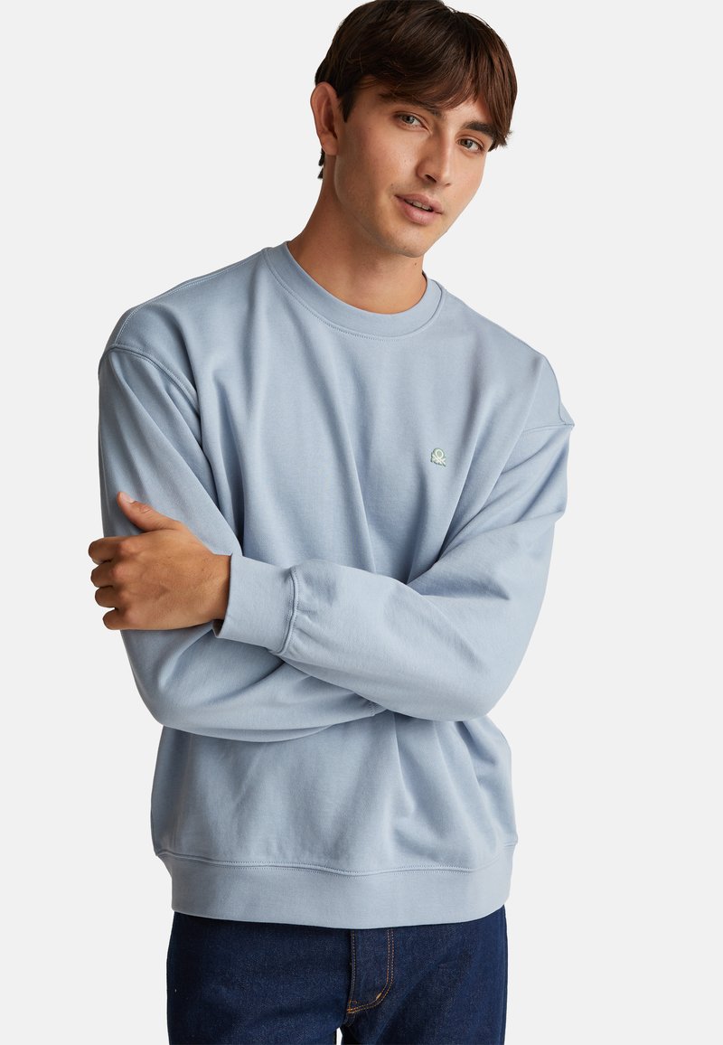 Sudadera de cuello redondo en azul claro, confeccionada en tejido suave. Presenta puños y dobladillo acanalados, con un pequeño logo verde en el pecho.