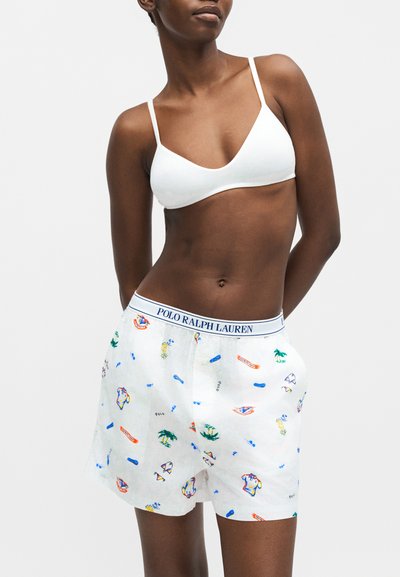 Γυναίκα που φορά λευκό bralette και λευκά boxer Polo Ralph Lauren με πολύχρωμα σχέδια, χέρια στις τσέπες, σε απλό φόντο.