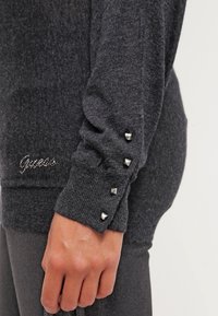Gros plan sur une manche tricotée gris foncé avec quatre clous pyramides argentés et un logo "Guess" en strass près du poignet sur le bras d'une personne.