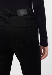 Personne portant un jean en denim noir avec une poche arrière visible et un patch en cuir de marque, vêtue d'un haut noir à manches longues, main détendue sur le côté.