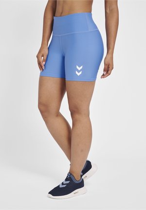 Kobieta w niebieskich sportowych spodenkach z wysokim stanem i granatowych wsuwanych butach sportowych z białymi logo w kształcie szewronu, stojąca na jednolitym tle.