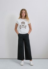 Camiseta blanca con una gráfica de calavera y huesos cruzados con estampado de leopardo, combinada con pantalones negros de pierna ancha y zapatillas blancas.